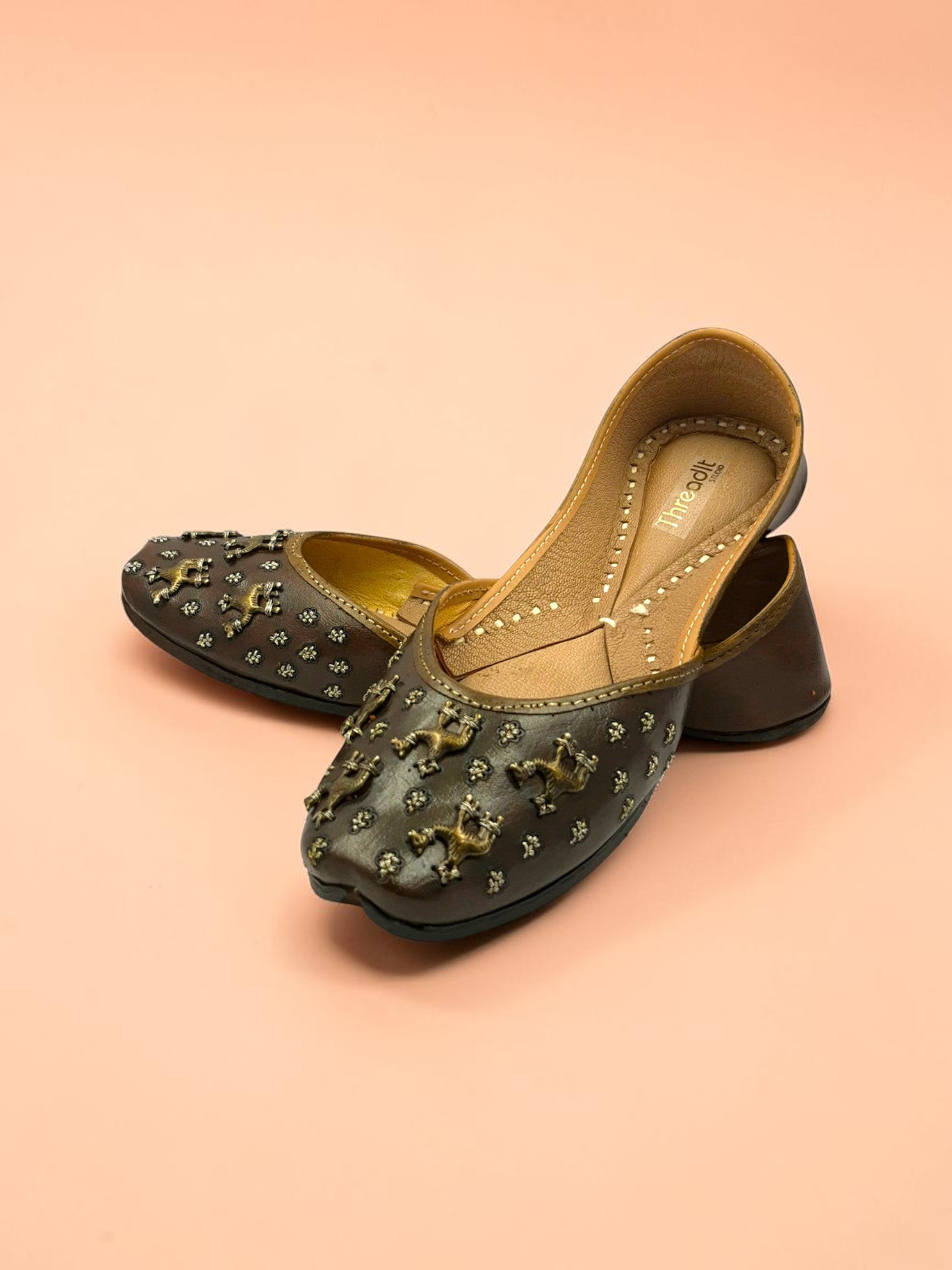 ThreadItStudio brown leather juttis with antique brass camel motifs and metallic studs on a soft peach background