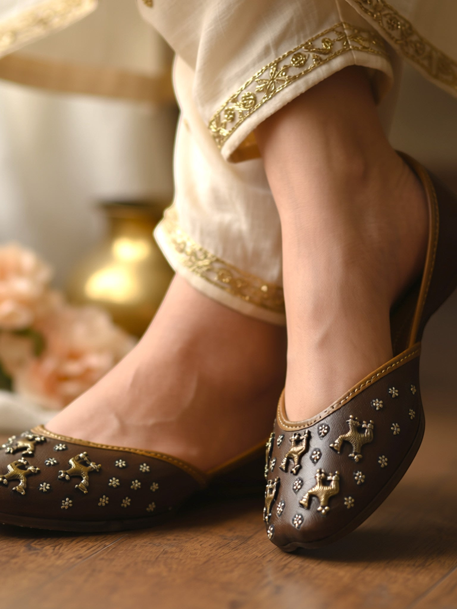 ThreadItStudio brown leather juttis featuring antique brass camel motifs and metallic studs worn with cream trousers.