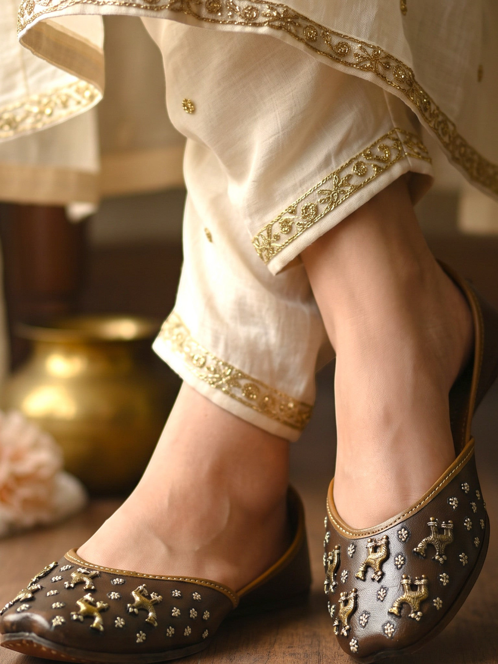 ThreadItStudio brown leather juttis with antique brass camel motifs and metallic studs worn with cream embroidered pants
