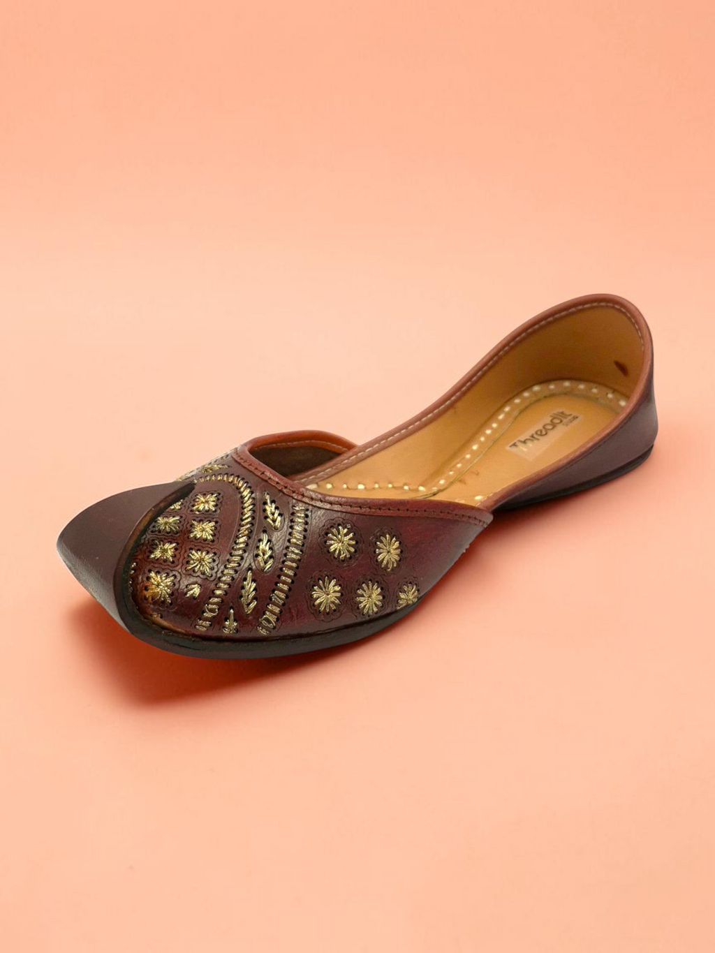 ThreadItStudio brown leather jutti with gold floral embroidery and geometric stitching on a peach background.