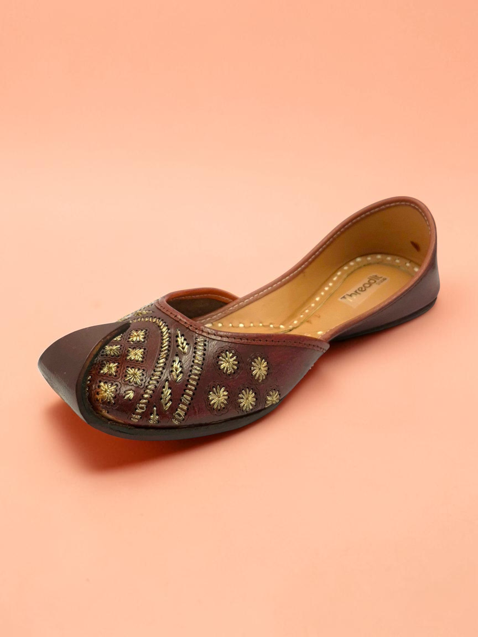 ThreadItStudio brown leather jutti with gold floral embroidery and geometric stitching on a peach background.